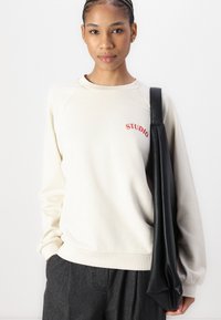 Sudadera crema con "STUDIO" en bordado rojo, cuello acanalado y corte relajado; combinada con pantalones gris oscuro y un bolso de piel negro.