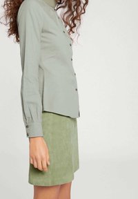 Vaaleanvihreä napitettava pitkähihaispaita, jossa on laskostetut kalvosimet, yhdistettynä samanväriseen mokkanahkaiseen minihameeseen. Yksinkertainen, klassinen muotoilu.