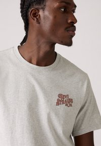 T-shirt de algodão cinza com um pequeno logótipo bordado em vermelho na área do lado esquerdo do peito, decote redondo e mangas curtas.