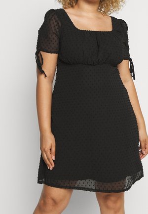Femme portant une robe noire, longueur genou, à manches courtes bouffantes et transparentes avec un motif à pois texturé, debout devant un fond uni.