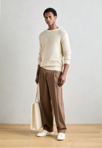 Pull en maille beige clair avec un motif texturé, pantalon marron ample, baskets blanches et sac fourre-tout crème. Fond en parquet en bois naturel.