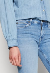 Jeans de mezclilla azul claro con un diseño ajustado, un suave desgaste y costuras en contraste. Combinados con una camisa de botones azul claro a juego.
