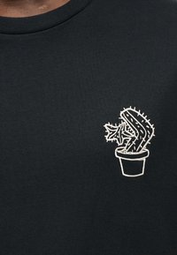 Μαύρο βαμβακερό μπλουζάκι με λευκό κεντητό σχέδιο κάκτου σε γλάστρα στην αριστερή πάνω πλευρά του στήθους. Ομαλή υφή και στρογγυλή λαιμόκοψη.