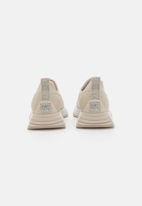 Zapatillas beige con parte superior texturizada y un logo reflectante de DKNY en el talón. Cuentan con una suela gruesa blanca y una pestaña de tira en la parte posterior.