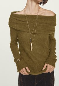 Olivgrüner, schulterfreier Pullover mit langen Ärmeln, weichem Stoff und gerippten Akzenten. Kombiniert mit geschichteten Halsketten und einem Armband.