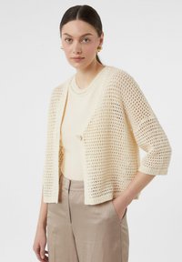 Strickjacke in Creme mit offenem Webdesign, gekürzten Ärmeln und einem einzelnen Knopfverschluss. Kombiniert mit einem beigen Oberteil und hellen Hosen.