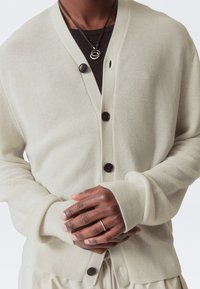 Cardigan beige clair avec un design en V, présentant des boutons foncés et des poignets côtelés. Porté par-dessus un vêtement noir, affichant un tricot texturé.