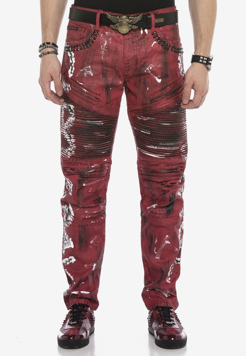 Rote Denim-Hosen mit distressed Akzenten, Reißverschlusstaschen und gerippter Textur. Verfügt über schwarze Nieten am Bund und dekorative Farbspritzer.