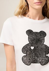 Witte T-shirt met een zwart kant teddybeer ontwerp met een open hart detail. Korte mouwen en een klassieke ronde halsstijl.