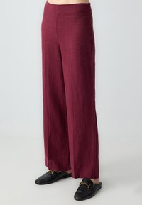 Wijde broek in diep bordeauxlinnen, met een gladde textuur en hoge tailleband. In combinatie met zwarte instappers met gouden accenten.