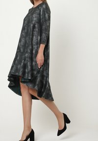 Femme portant une robe asymétrique gris délavé à manches longues avec un ourlet à volants, associée à des chaussures noires à talons blocs.