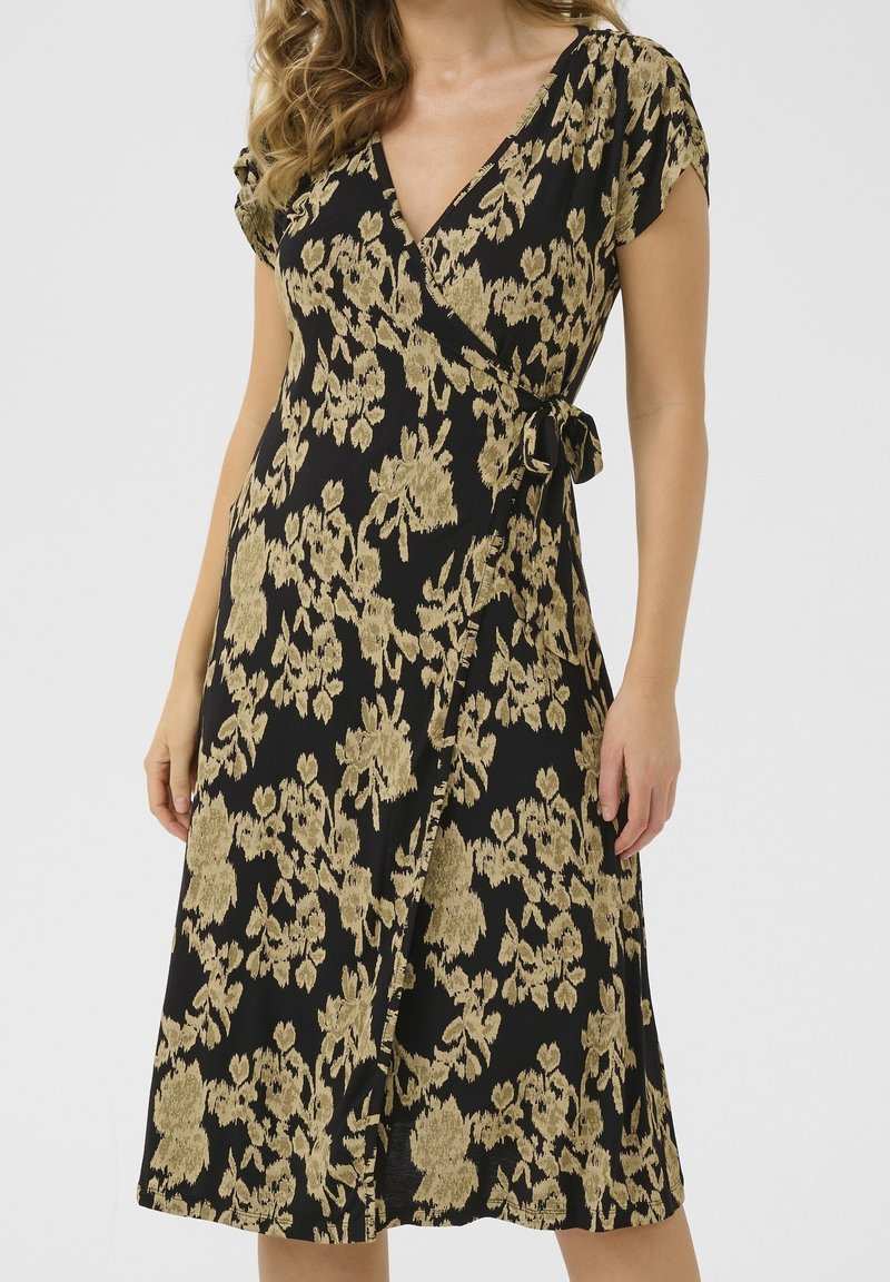 Femme portant une robe portefeuille noire avec un motif floral beige, manches courtes, col en V et détail de nouage sur le côté.