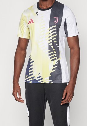 Homme portant un maillot de sport à manches courtes avec un motif abstrait jaune, noir et blanc et un pantalon de sport noir avec des rayures blanches.