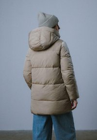 Beżowa kurtka puchowa z kapturem, pikowana, wykonana z mocnego materiału. W połączeniu z szarym, prążkowanym czapkiem i jasnoniebieskimi dżinsami.