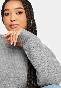 Grå stickad tröja med rund halsringning och långa ärmar, med ett texturerat mönster. Flätat hår och naturliga naglar syns i förgrunden.