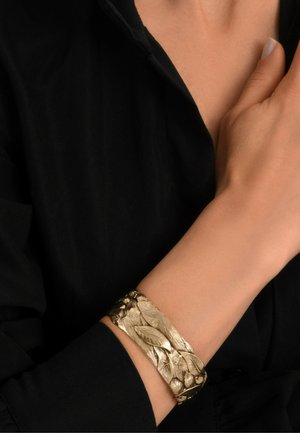 Bracelet jonc doré à l'allure texturée en forme de feuille ; large et lisse avec des accents de feuilles en relief, porté sur un poignet nu.