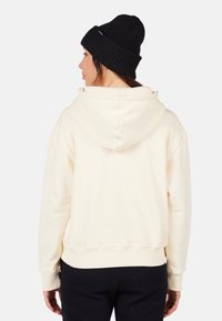 Krémová mikina s kapucí na šňůrku, ribbovaný lem, pohodlný střih, nošena s černou pletenou baretou. Zadní pohled ukazuje jednoduchý design.