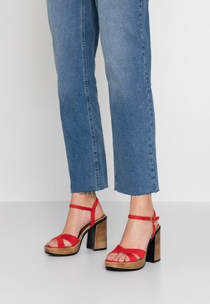 Benen met blauwe cropped jeans en rode high-heeled sandalen met houten plateau en enkelbandjes op een witte vloer achtergrond.