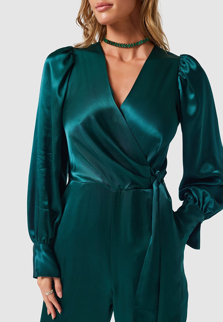 Frau in einem smaragdgrünen satinartigen Wickel-Jumpsuit mit gepufften langen Ärmeln, tiefem V-Ausschnitt und einem mit Juwelen besetzten Halsband, die eine Hand in der Tasche hat.