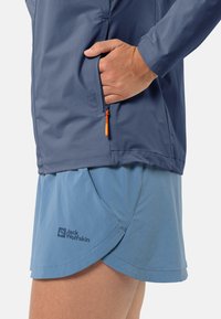 Blauwe lichte jas met oranje ritsaccenten en een zak. Gecombineerd met blauwe, gestructureerde shorts met een logo op de onderste zoom.