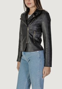 Giacca da biker in pelle nera con design aderente, tasche con zip e colletto a punta. Texture liscia e dettagli in metallo.