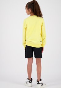 Felpa gialla con polsini a costine, abbinata a pantaloni shorts neri. Le calzature presentano dettagli bianchi con stelle nere. Vista da dietro.