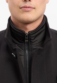Schwarze Jacke mit hohem Kragen, strukturierter Oberfläche, Reißverschluss vorne und gestepptem Innenfutter. Mit Druckknopfdetail am Kragen.