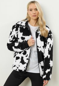 Veste en polaire à motif de vache noir et blanc avec une fermeture éclair à l'avant, deux poches et une texture douce. Portée sur une chemise blanche et un legging noir.