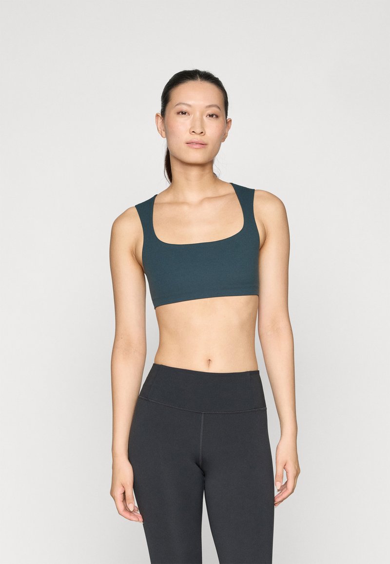 Sujeción deportiva en color verde azulado con tirantes anchos y escote en forma de bote, confeccionada con un material suave y elástico. Combinada con leggings negros de cintura alta.