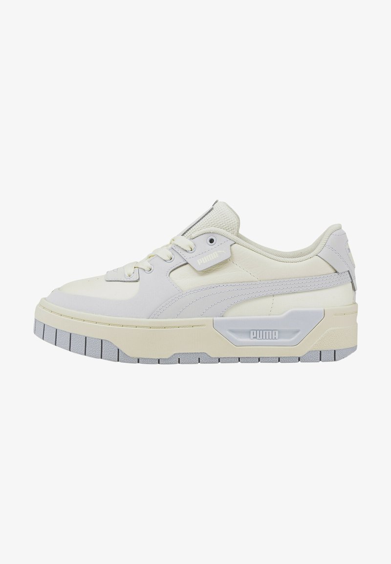 Light beige sneaker with a textured upper, lace-up design, and a thick grey sole. Features a prominent logo accent on the side.