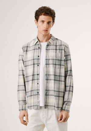 Giovane uomo con capelli corti e ricci che indossa una camicia a quadri grigio chiaro e bianca sopra una t-shirt bianca e pantaloni chiari.