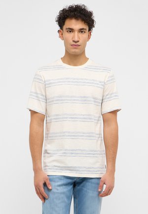Junger Mann mit lockigem Haar, der ein cremefarbenes T-Shirt mit hellblauen horizontalen Streifen und hellblaue Jeans trägt, stehend vor einem schlichten Hintergrund.