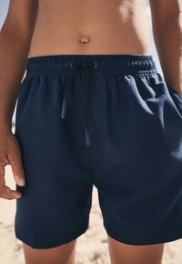 Tmavomodré plavecké šortky s elastickým pásom a čiernym sťahovacím šnúrkou. Jemná tkanina s bočnými vreckami a pohodlným strihom.