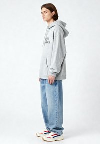 Szürke pamut kapucnis pulóver kenguruzsebbel, hímzett szöveggel a mellkasán, világoskék bő szabású farmernadrággal és fehér edzőcipővel, piros részletekkel.