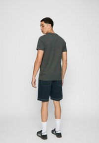Tricou gri din bumbac cu gât rotund, asociat cu șorturi bleumarin cu cusături albe pe buzunare. Modelul stă în picioare, cu spatele la cameră.