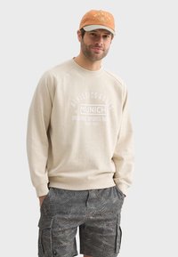 Sweat-shirt beige avec impression blanche, arborant la marque "Munich" ; associé à un short cargo gris à motifs ; le modèle porte une casquette orange.