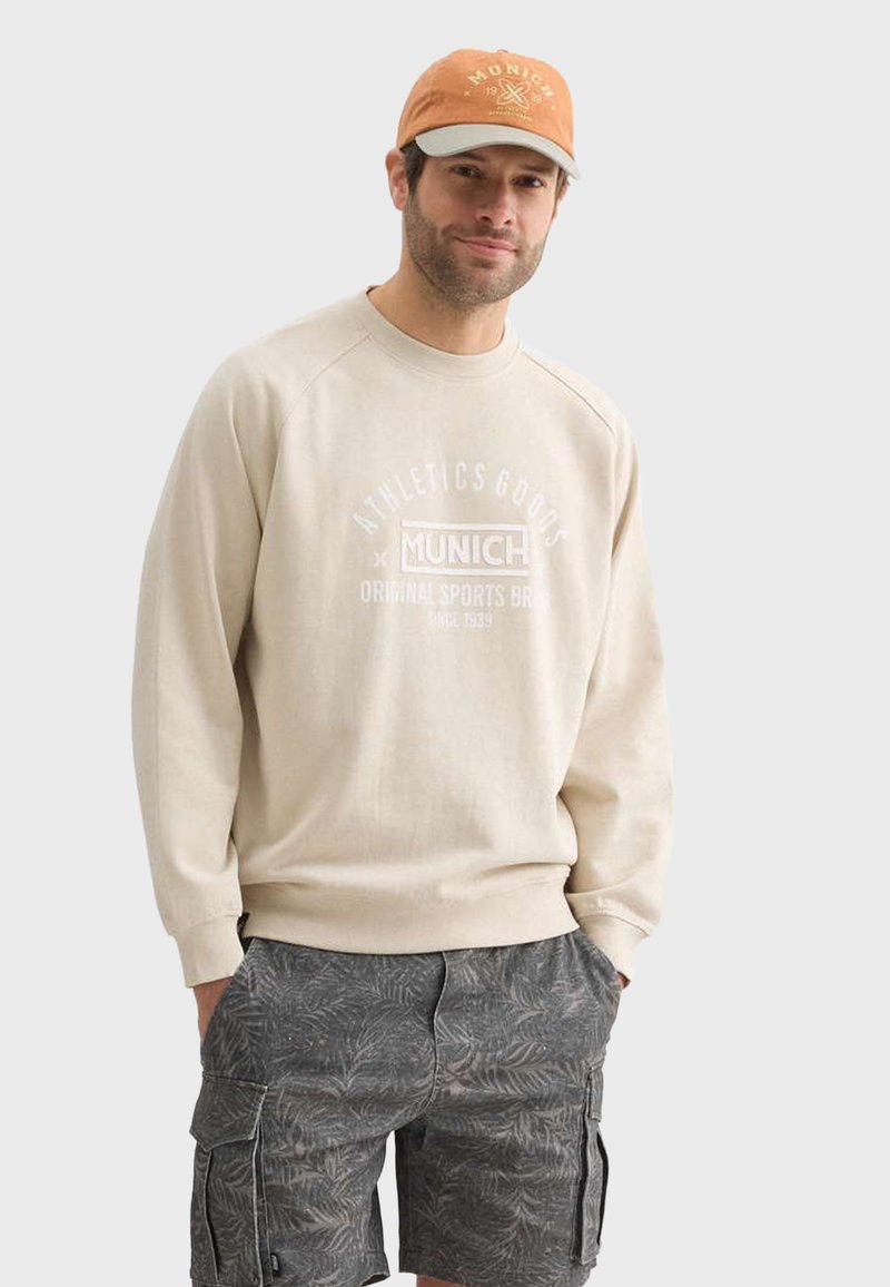 Sweat-shirt beige avec impression blanche, arborant la marque "Munich" ; associé à un short cargo gris à motifs ; le modèle porte une casquette orange.