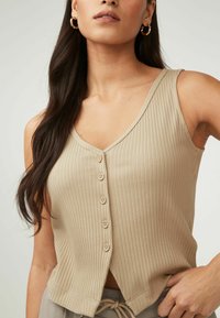 Top sin mangas de canalé beige con escote en V, que presenta seis botones decorativos en la parte delantera y un ajuste ligeramente acampanado en el dobladillo.