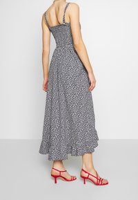 Kvinna i en svart och vit blommig midi-klänning med tunna band och röda klacksandaler, stående på en grå golv mot en vit vägg.