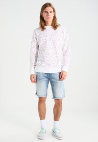 Felpa bianca con un motivo a pois multicolori, polsini a coste e scollatura rotonda, abbinata a pantaloni corti in denim blu chiaro e sneakers.