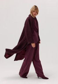 Przesadnie duży bordowy płaszcz z teksturowanej tkaniny, w zestawieniu z dopasowanym garniturem i szpilkami o spiczastych noskach, na neutralnym tle.