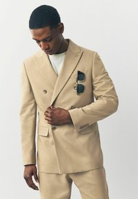 Abito beige a doppio petto realizzato in tessuto liscio, con silhouette su misura, due tasche frontali e occhiali da sole nella tasca del petto.