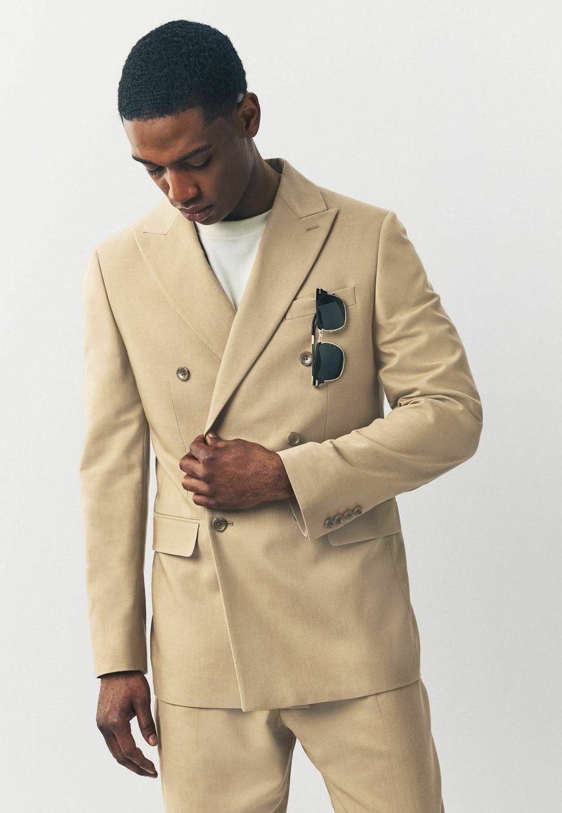 Abito beige a doppio petto realizzato in tessuto liscio, con silhouette su misura, due tasche frontali e occhiali da sole nella tasca del petto.