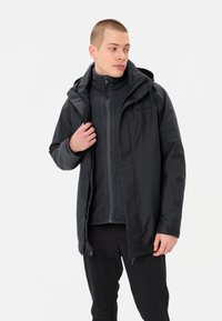 Giacca nera impermeabile con cappuccio, caratterizzata da un design elegante e una vestibilità affusolata. Sotto, è visibile un maglione zip-up testurizzato scuro.