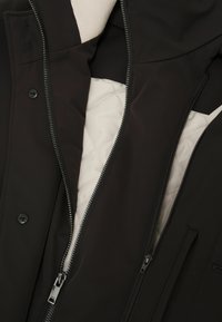 Schwarze Jacke mit zwei vorderen Reißverschlüssen, teilweise geöffnet, die ein gesteppte hellbeige Innenfutter und darunter einen cremefarbenen Rippstoff sichtbar machen.
