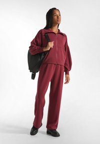 Completo tuta color bordeaux in morbido tessuto con giacca con zip e pantaloni larghi. Zaino nero con spallacci e scarpe nere piatte.