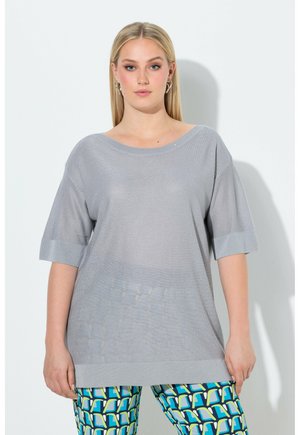 Frau trägt ein lockeres, hellgraues Kurzarmoberteil und Leggings mit geometrischem Muster in Blau, Grün und Weiß, steht vor einer weißen Wand.