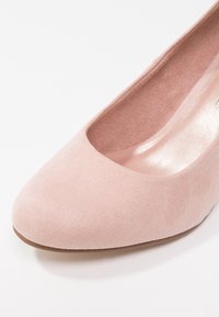 Gros plan sur une chaussure plate en daim rose clair, à bout arrondi et semelle intérieure douce, sur fond blanc.