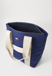 Mörkblå canvas axelväska med beige handtag, utrustad med dragkedja upptill och en märkesbricka på framsidan för identifiering.