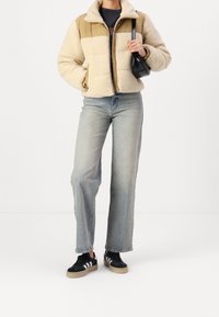 Kremowo-beżowa kurtka polarowa na zamek, o luźnym kroju, uzupełniona jasnoniebieskimi, prostymi jeansami i czarnymi sneakersami.